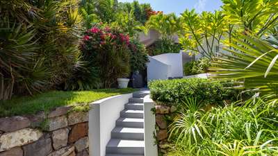 Elegant grey steps leading to the contemporary white exterior of Villa Adharra, framed by vibrant tropical gardens.
