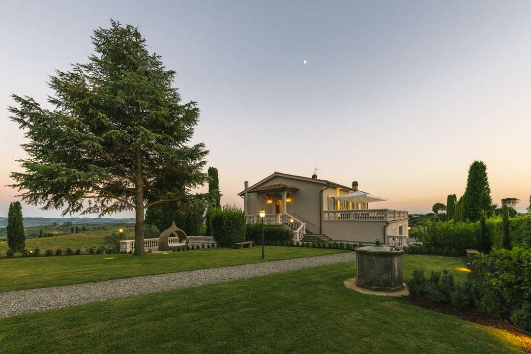 Elegant XV century Tuscan villa exterior at dusk with a manicured lawn, gravel path, and sweeping views of rolling hills.