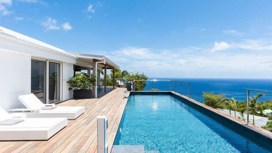 Expansive wooden terrace with plush white sunbeds beside a 4x12m private infinity pool, offering panoramic ocean views.