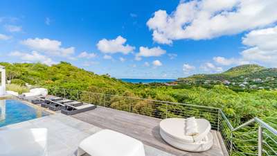 Expansive wooden terrace featuring a modern white daybed, infinity pool, and sweeping ocean views over St. Jean bay.