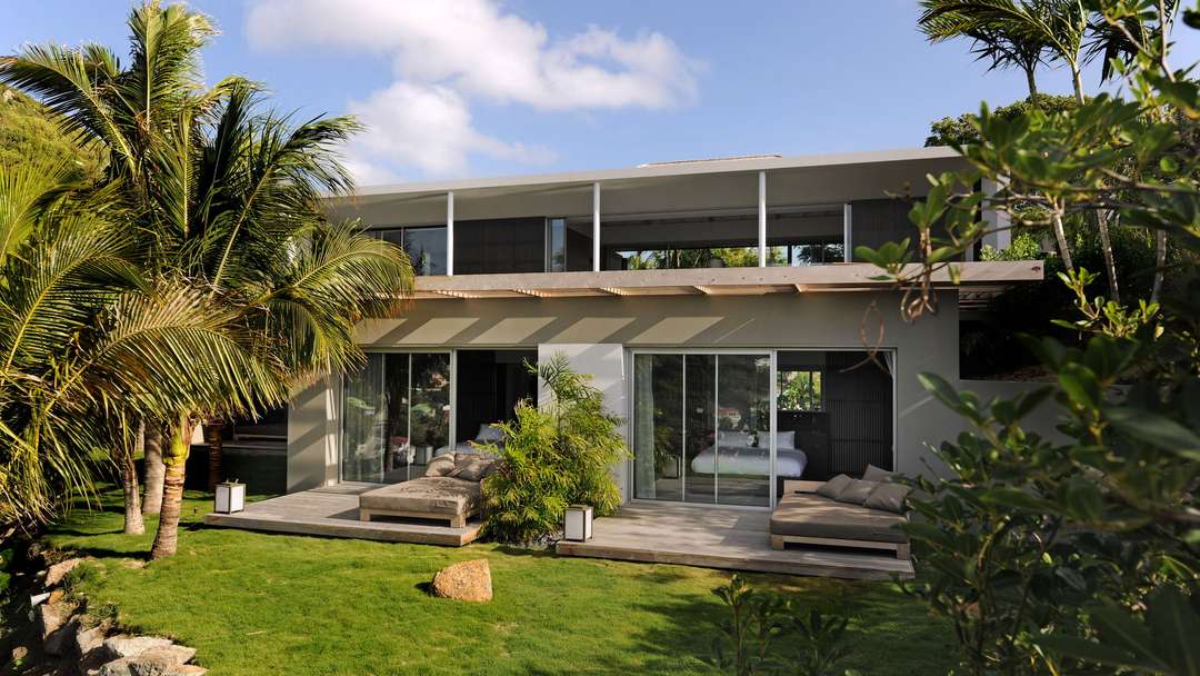 Lower-level bedrooms at Villa Bakea open to a wooden deck with plush sun loungers, surrounded by lush tropical gardens.
