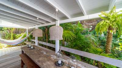 Covered alfresco dining area at Villa Bibi with a rustic wood table, woven pendants, and a hammock amidst tropical gardens.