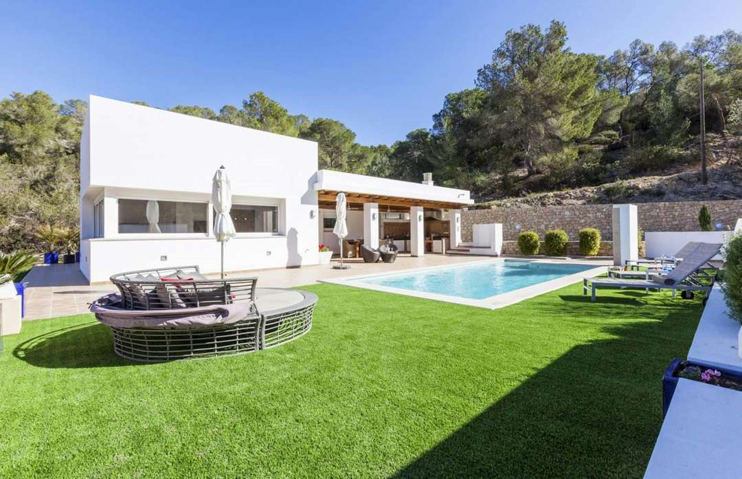 Elegant white villa exterior featuring a private pool, plush daybed, and lush green lawns at By the Sands in Cala Vadella.