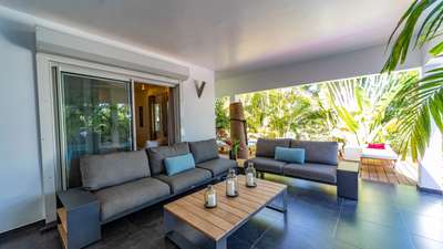 Chic covered outdoor salon featuring comfortable grey sofas and a wooden coffee table, opening to the sunlit pool deck.