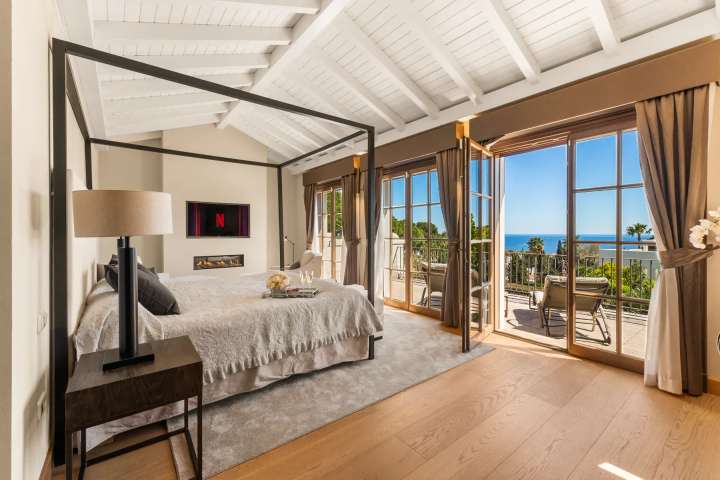 Master bedroom with a four-poster bed, modern fireplace, and private balcony offering stunning Mediterranean Sea views.