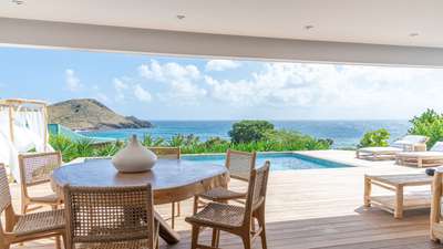 Elegant alfresco dining area on a covered terrace, featuring a natural wood table, woven chairs, and breathtaking ocean views.