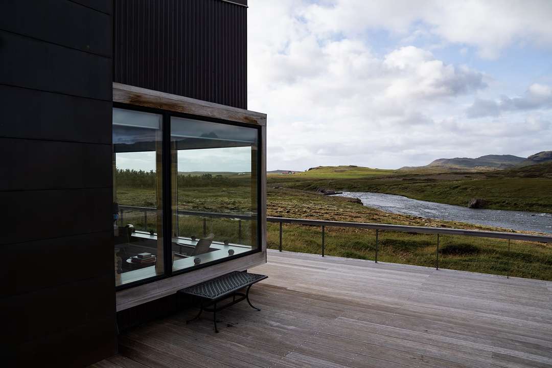 Modern luxury lodge exterior with dark cladding, expansive glass windows, and a wooden deck overlooking Iceland's scenic river.