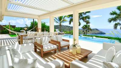 Sun-drenched outdoor lounge under an adjustable pergola, featuring teak furniture and an infinity pool merging with the sea.