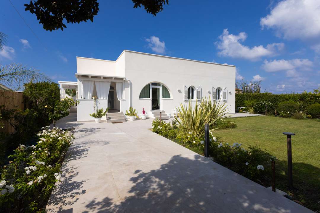 Elegant white waterfront villa in Sicily featuring classic arched windows, a paved pathway, and lush Mediterranean gardens.