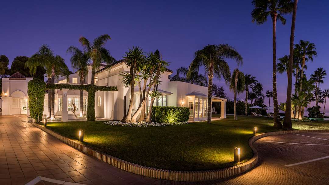 Villa Miro's elegant white exterior illuminated at dusk, featuring manicured lawns and towering palms in Los Monteros.