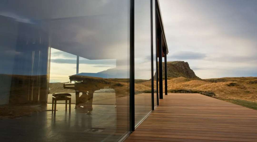 Grand piano visible through floor-to-ceiling windows at Fish Creek, reflecting Borgarfjörður's rugged mountain landscape.