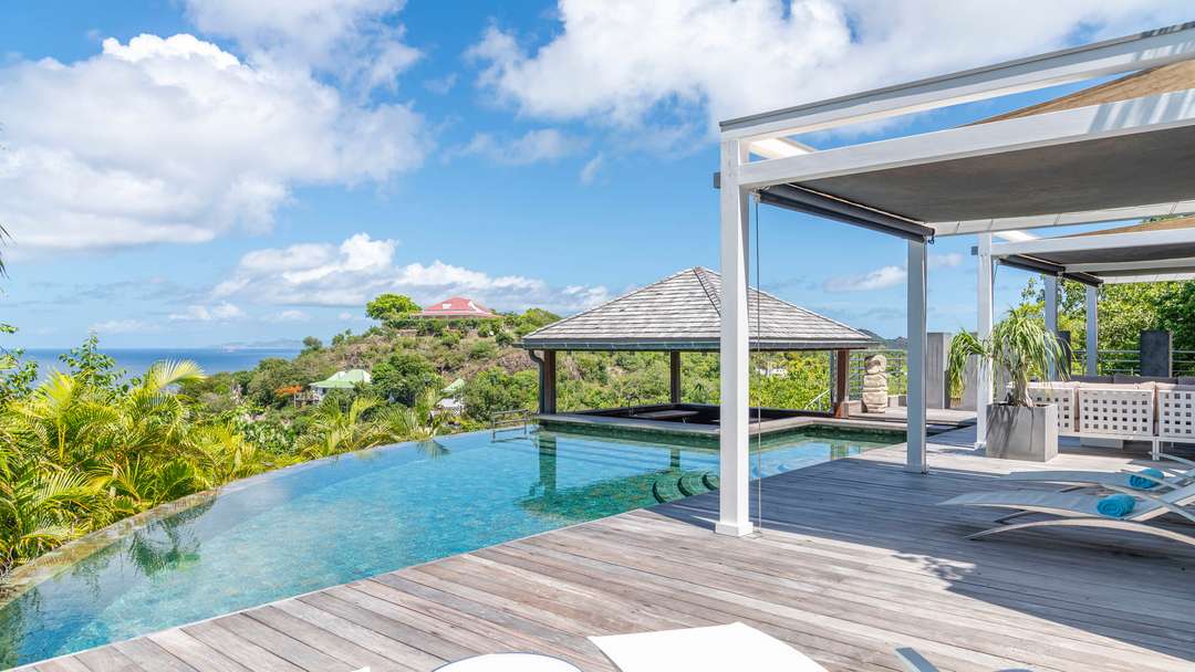Heated infinity pool and wooden terrace at Villa Fleur De Cactus, featuring a gazebo, wet bar, and stunning ocean views.