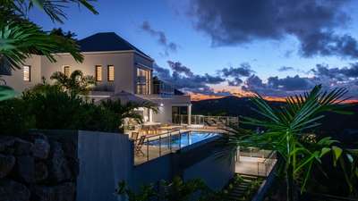 Stunning multi-level luxury villa illuminated at dusk, featuring an infinity pool, wooden decks, and tropical gardens.