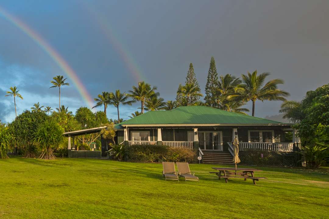 Beachfront Hanalei Bay Hale exterior with green roof, lush lawn, palm trees, and a stunning double rainbow in Kauai.