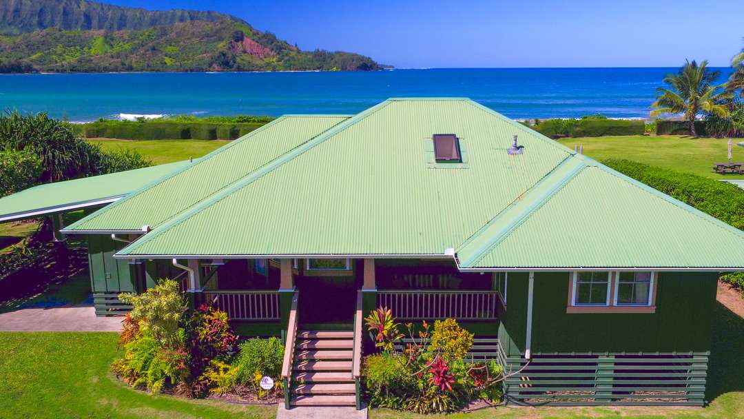Hanalei Nalu House aerial view showcasing its olive-green roof, beachfront setting, and stunning Kauai mountain backdrop.