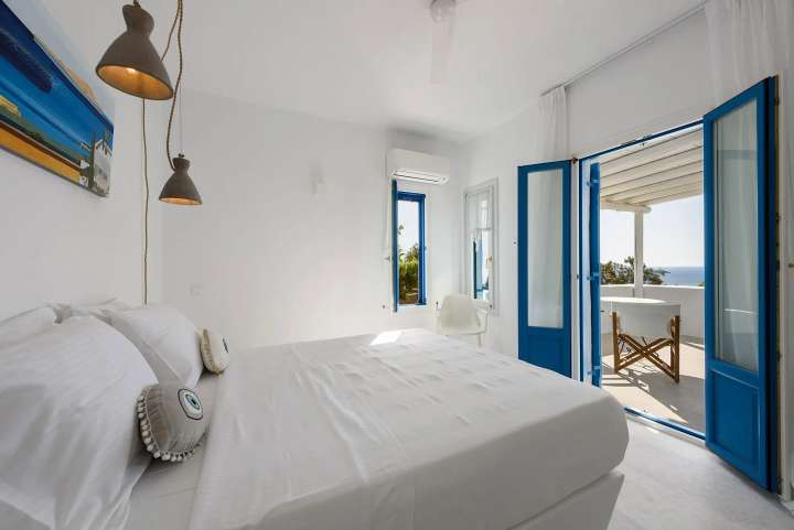 Elegant double bedroom in a luxury Cycladic villa with blue veranda doors opening to a balcony and Aegean Sea views.