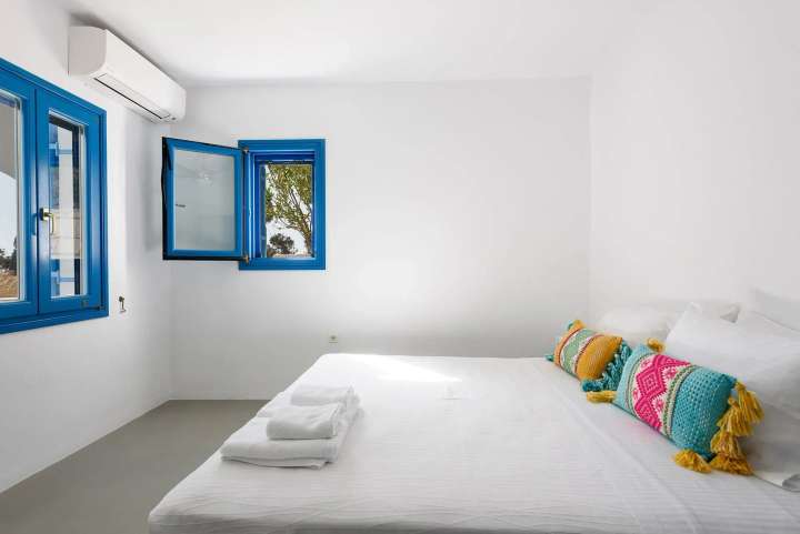 Minimalist whitewashed double bedroom at Hidden Jasmine, featuring traditional blue windows and vibrant accent pillows.