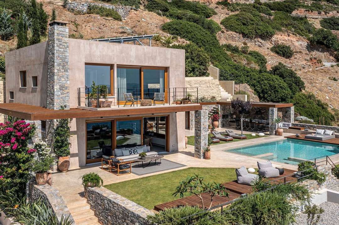 Luxury Heraklion villa exterior with heated infinity pool, stone facade, and elegant outdoor lounge against a hillside backdrop.