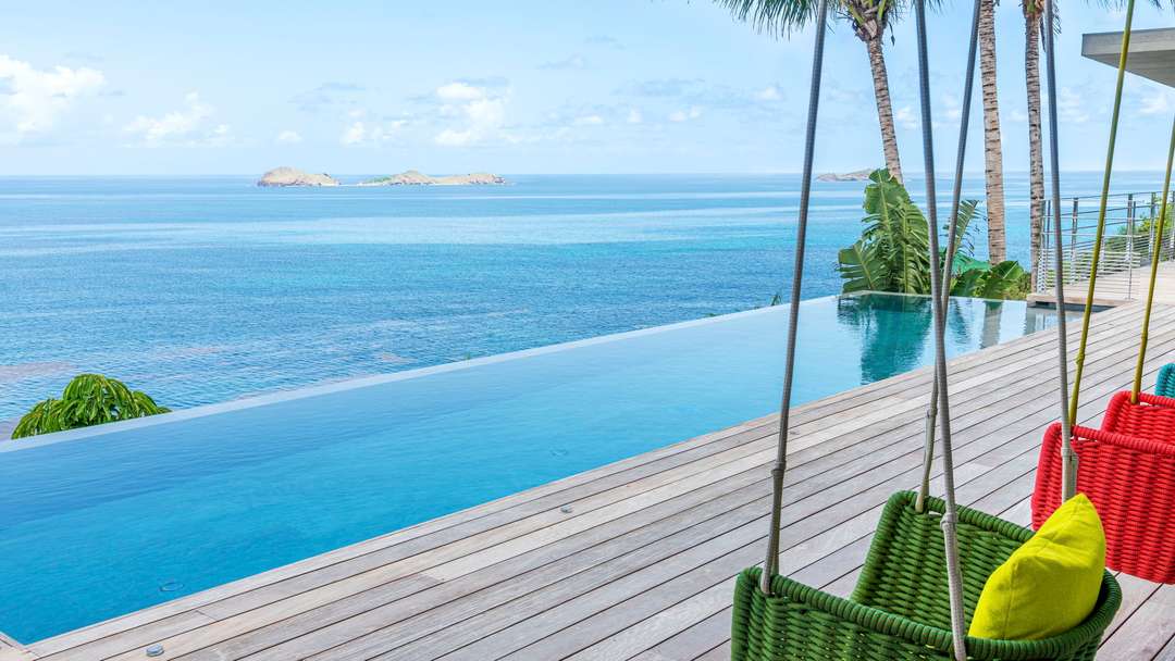 Heated infinity pool and wooden deck with colorful hanging chairs overlooking panoramic ocean views at Villa Honu, St-Barths.