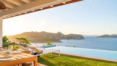 Sun-drenched terrace with plush lounge chairs beside a shimmering infinity pool, offering panoramic St-Jean bay views.