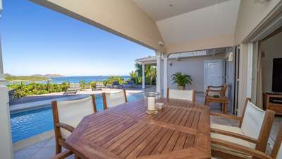 Elegant outdoor dining area under a covered terrace, featuring a wooden table and panoramic views of the Caribbean bay.