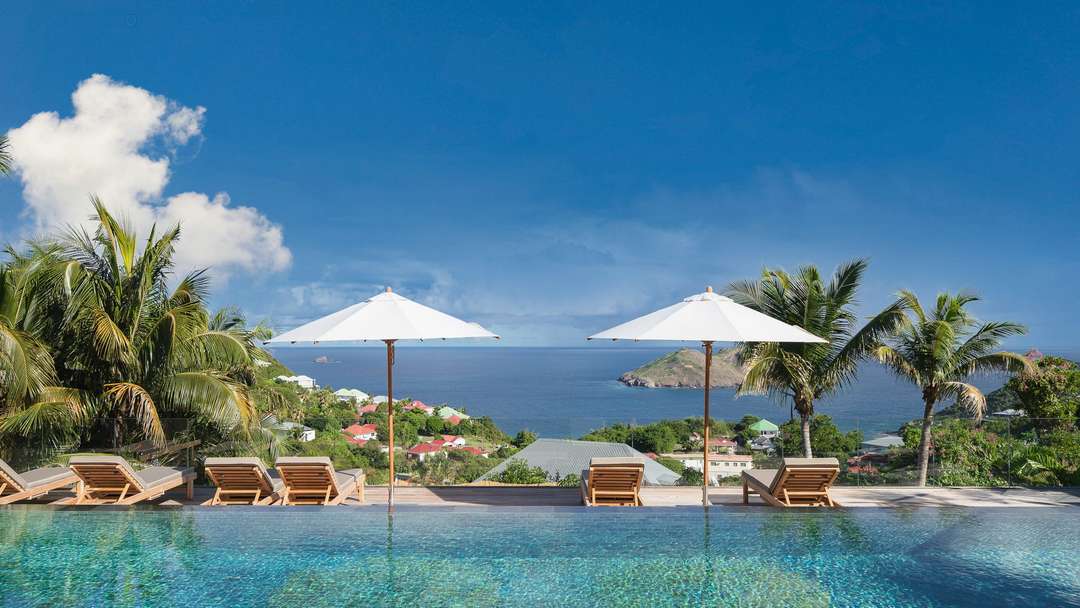 Heated infinity pool and wooden terrace with sun loungers at Villa Les Palmes, offering panoramic ocean views of Flamands Bay.