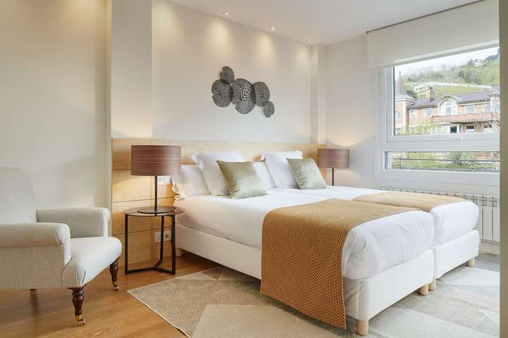 Elegant en-suite bedroom offering twin beds, light wood headboard, and large windows with peaceful San Sebastián town views.