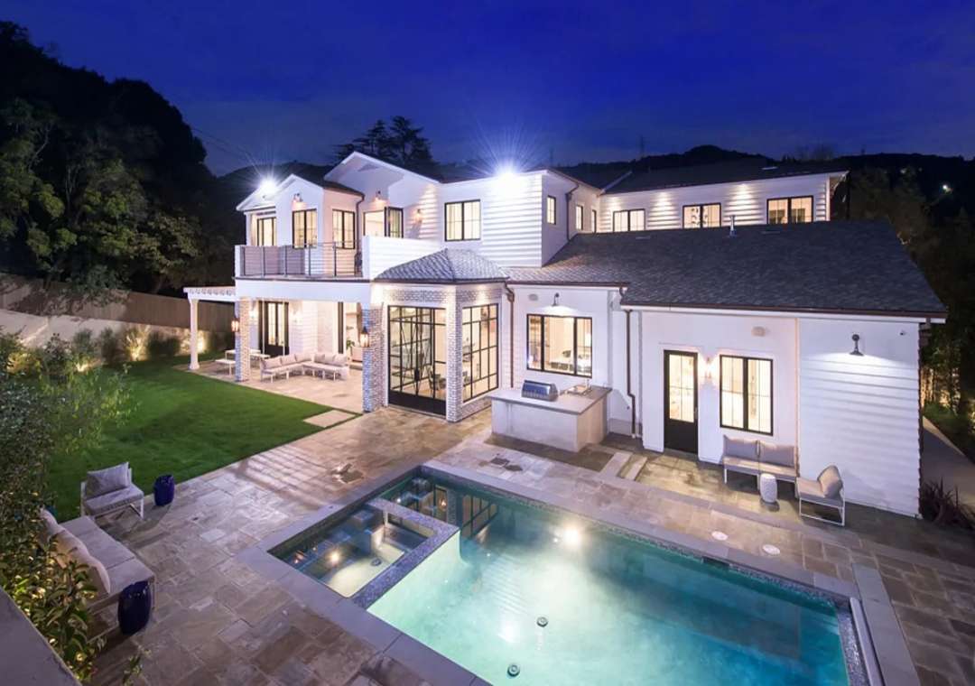 Luxury Los Angeles contemporary farmhouse exterior at night, featuring a sparkling pool, spa, and expansive grassy lawn.
