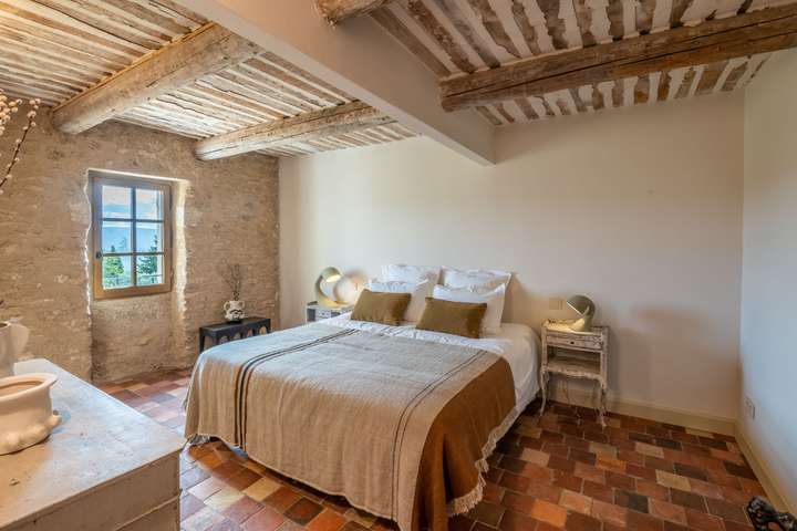 Elegant bedroom at Mas des Altesses featuring exposed beams, stone walls, terracotta floors, and scenic Gordes village views.
