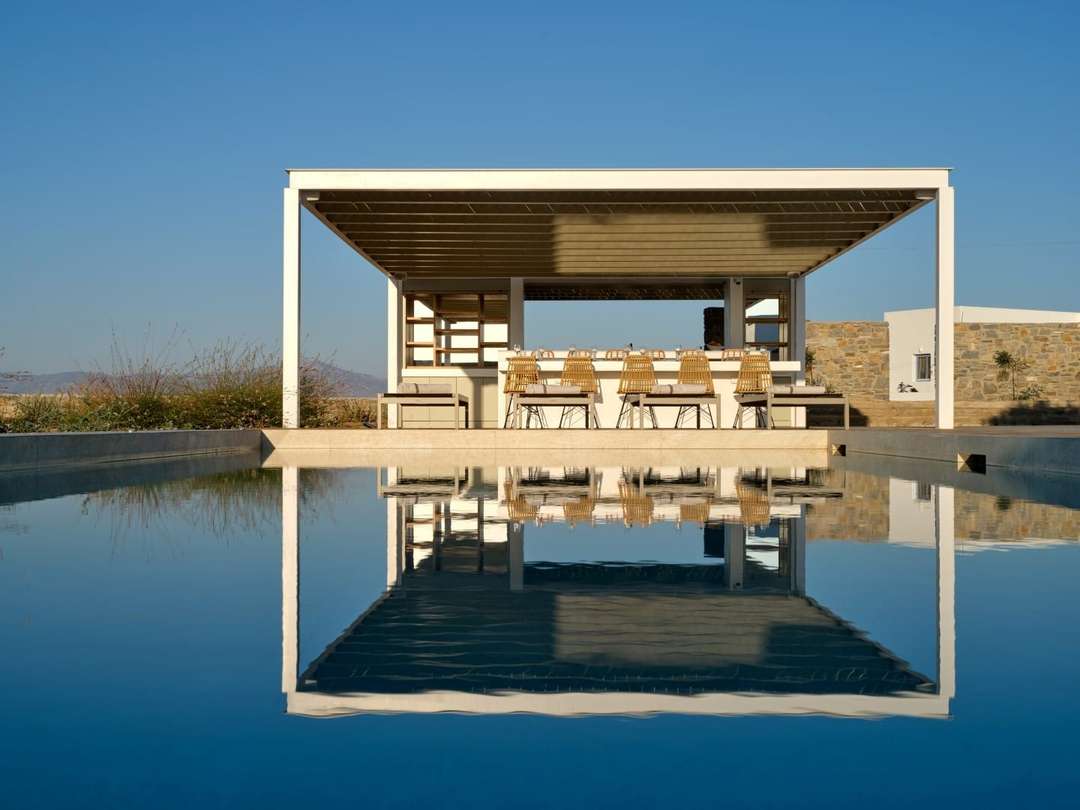 Elegant outdoor bar and dining area under a white pergola, beautifully reflected in the private pool at Naousa Tide villa.