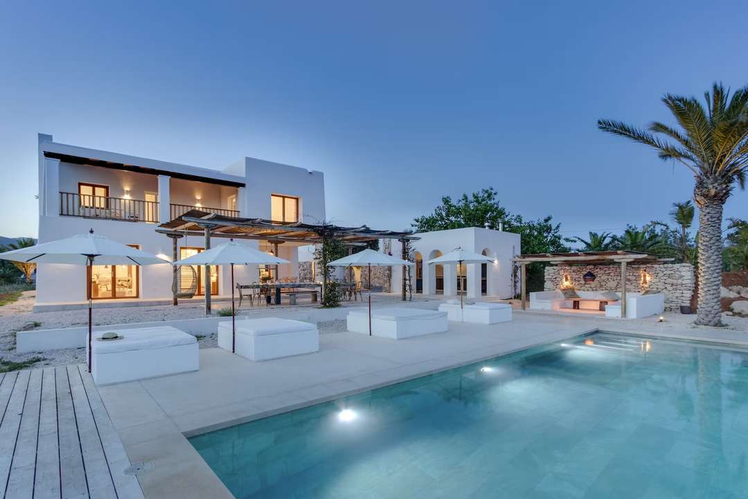 Elegant whitewashed Ibiza villa at dusk featuring a 12m private pool, luxurious daybeds, and wooden-beamed verandas.