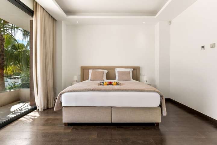 Elegant en-suite bedroom featuring rich dark wood flooring, a plush bed, and sliding glass doors leading to a sunny balcony.