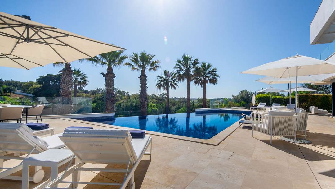 Elegant sun loungers beside a pristine infinity pool with palm trees and lush gardens at this high-end Algarve villa.