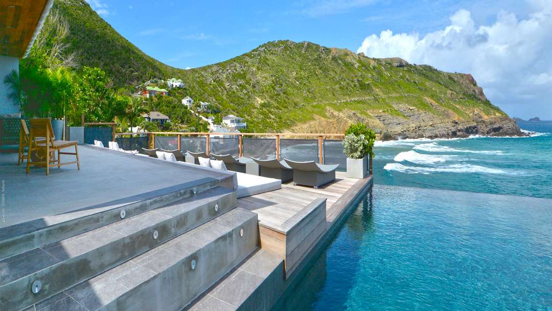 Heated infinity pool and wooden terrace with sun loungers overlooking the ocean and lush hills of St. Barths.