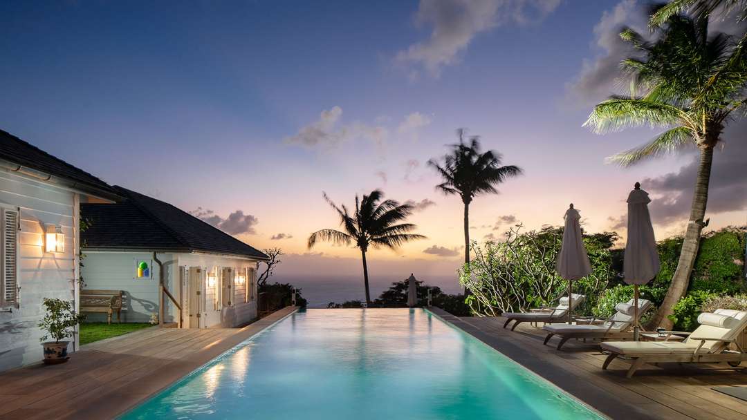 Illuminated 17-meter heated pool at dusk at Villa Plumbago, featuring a wooden deck, white shutters, and Caribbean Sea views.