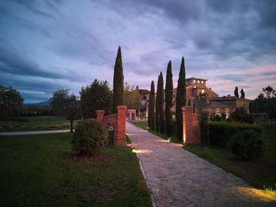 Illuminated stone pathway with tall cypress trees leading to the historic Poggio Prezioso resort at twilight in Tuscany.