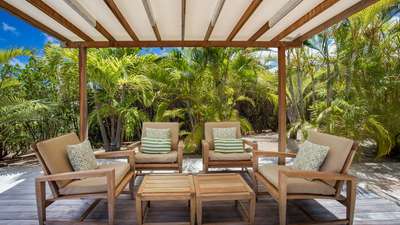 Shaded outdoor lounge beneath a wooden pergola, featuring elegant teak armchairs surrounded by lush, swaying palm trees.