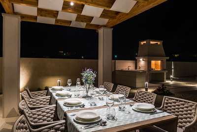 Alfresco dining terrace under a wooden pergola, seating eight, with a traditional stone barbecue grill illuminated at night.