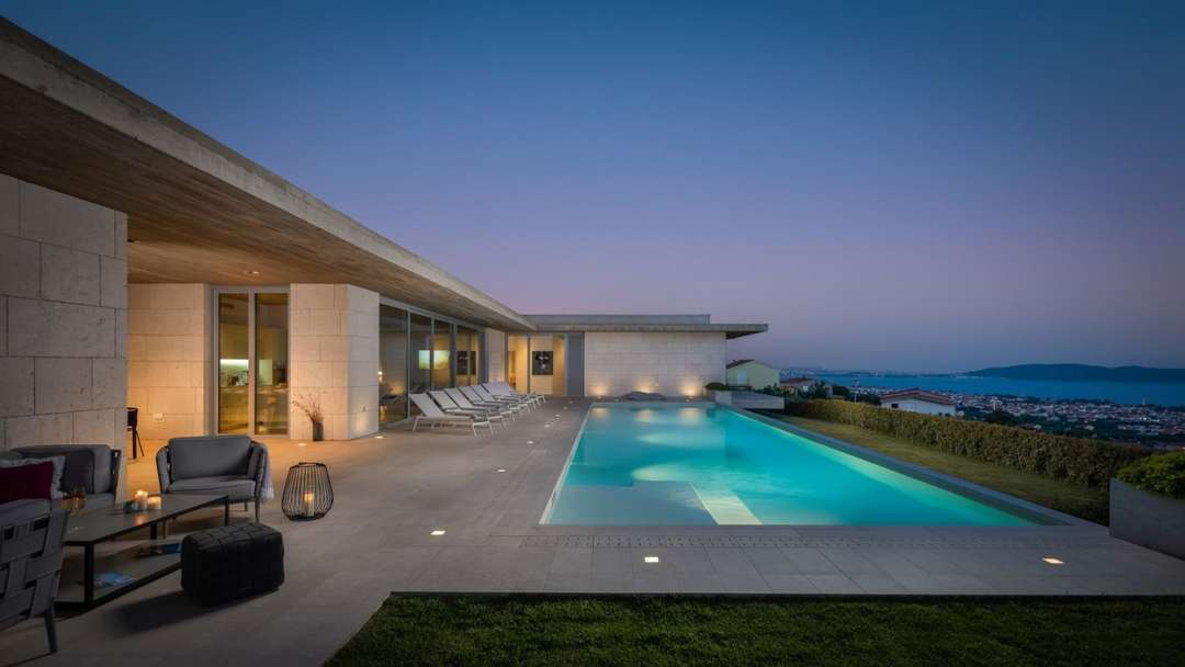 Modern luxury villa terrace at dusk featuring a heated infinity pool, elegant loungers, and panoramic sea views over Split.