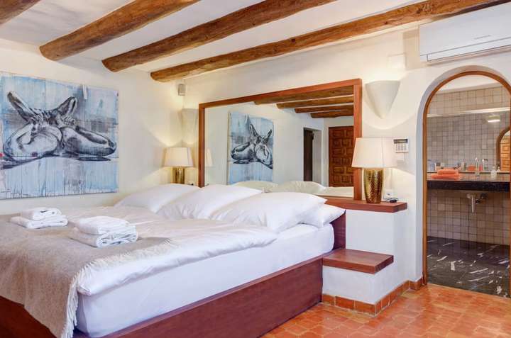 Master suite featuring a king-size bed, natural wood beams, terracotta floors, and direct access to an en-suite bathroom.