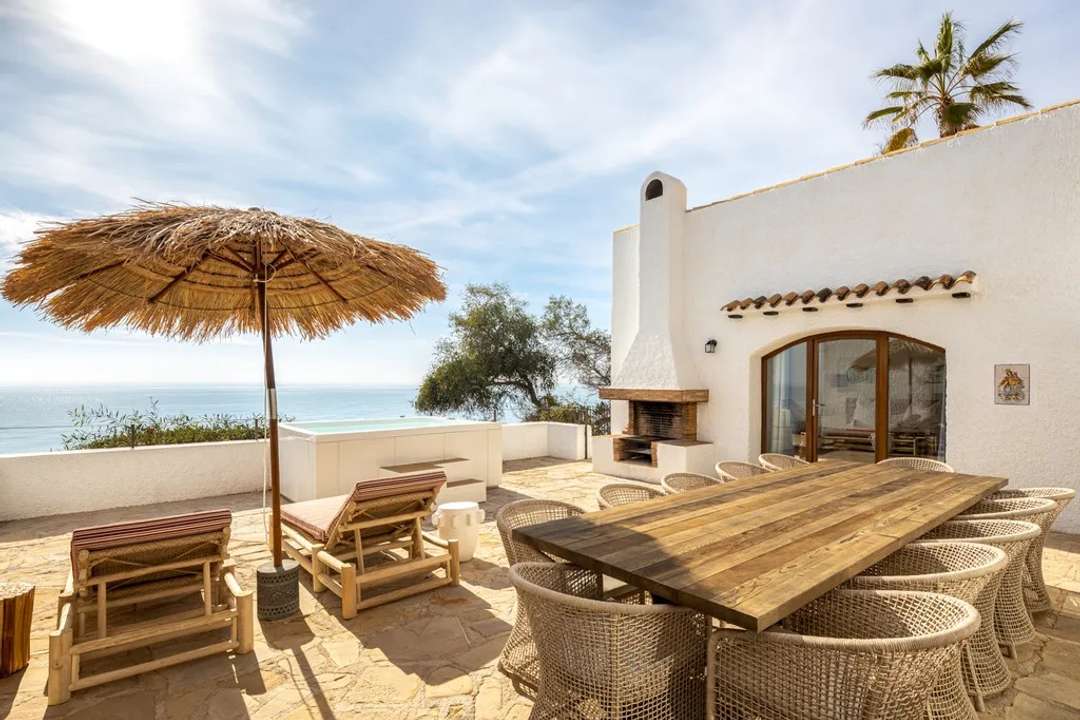 Expansive stone terrace at Sunset Staircase featuring a wooden dining table, waterfront jacuzzi, and outdoor fireplace.