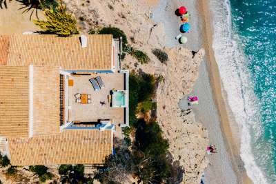 Aerial view of a luxury stone terrace featuring a waterfront jacuzzi, outdoor dining, and direct beach access below.