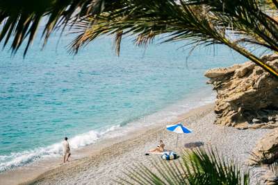 Exclusive pebble beach with crystal-clear Mediterranean waters and dramatic rock formations, framed by lush palm fronds.
