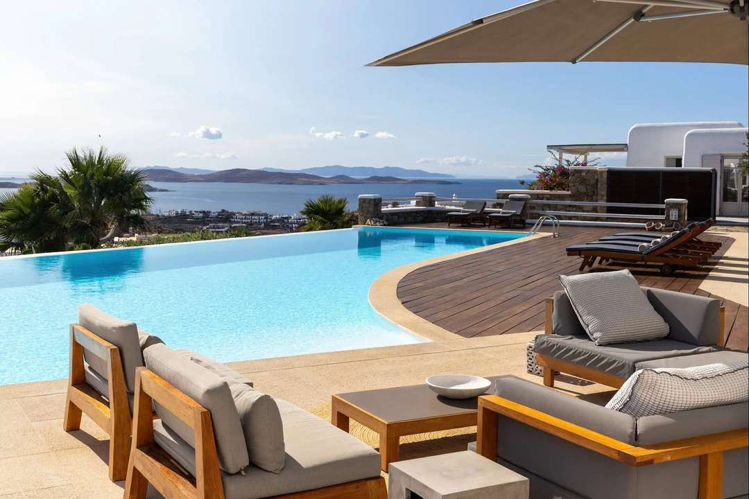 Luxury infinity pool and wooden deck at Villa Alchemia, featuring sun loungers and panoramic Aegean Sea views in Mykonos.