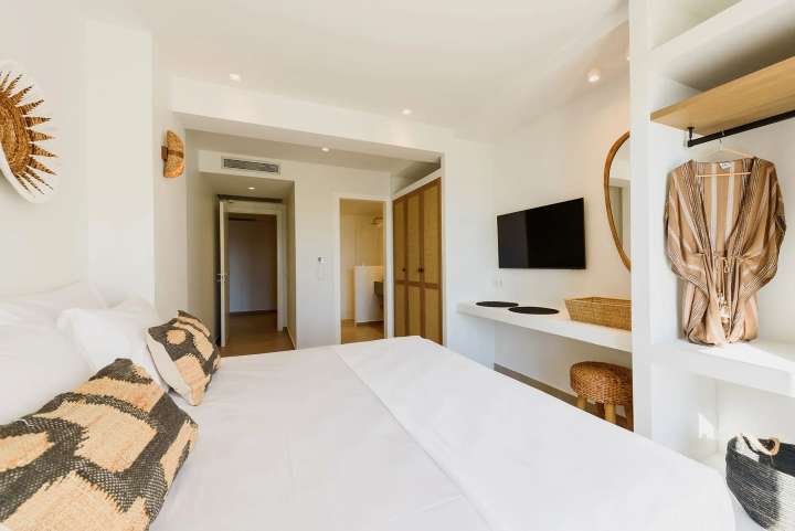Luxurious double en-suite bedroom with woven accents, a flat-screen TV, and custom open closets in a modern Cycladic style.