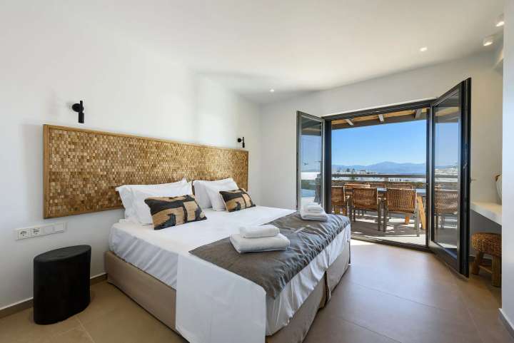 Elegant double bedroom with custom natural textures and large glass doors opening to a shaded terrace with sweeping sea views.