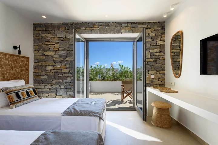 Stylish twin bedroom featuring a Cycladic stone wall and large sliding glass doors opening to a private sea view terrace.