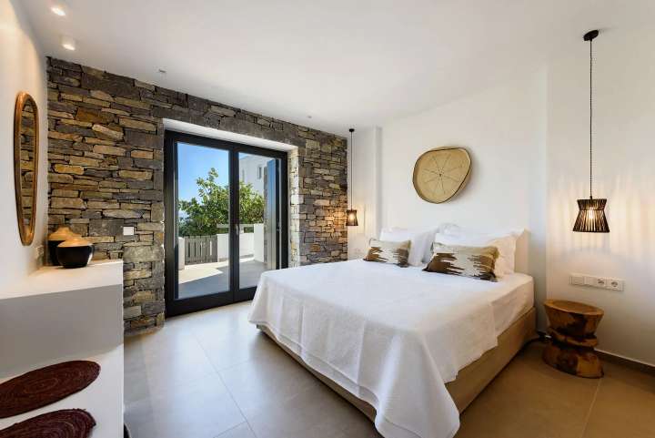 Luxurious double bedroom blending Cycladic stone walls with modern design, opening to a terrace with beautiful sea views.