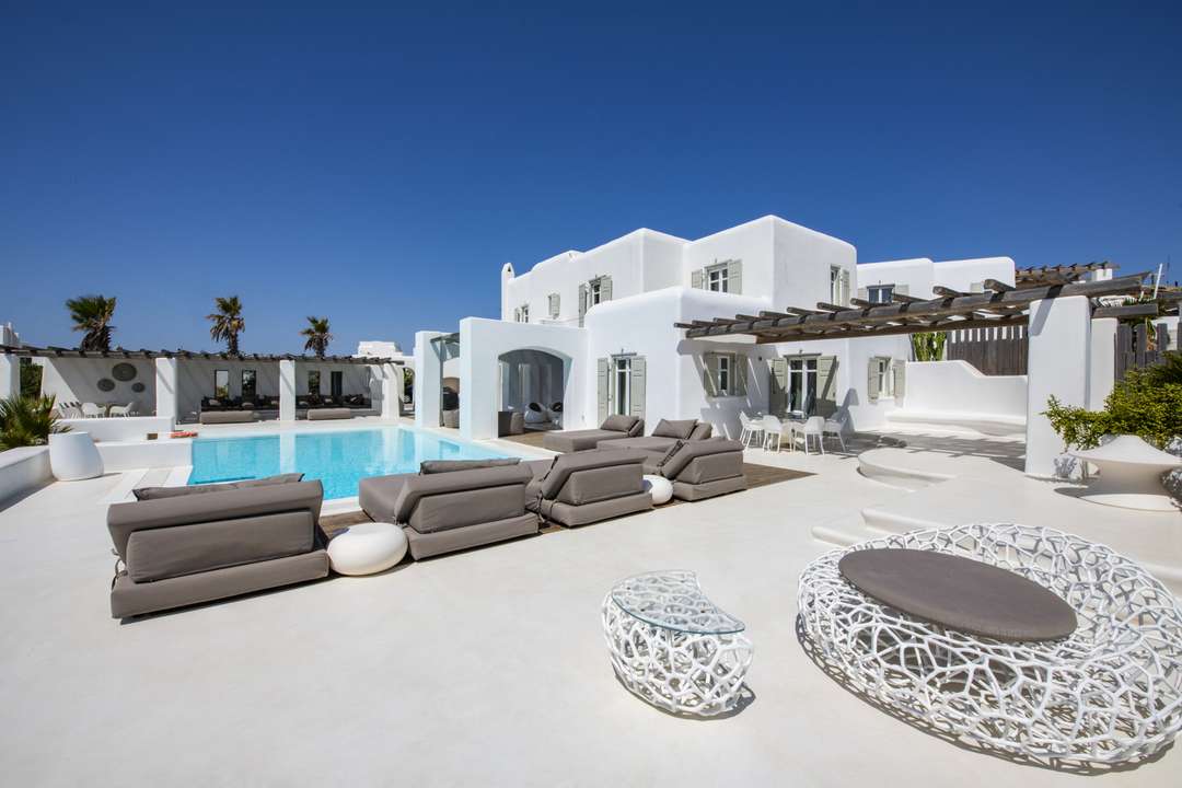 Luxury Mykonos villa with whitewashed exterior, infinity pool, modern loungers, and shaded wooden pergolas for relaxation.