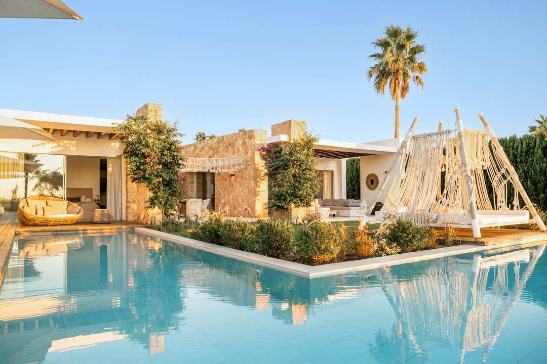 Luxury Ibiza villa with L-shaped infinity pool, natural stone walls, and a boho-chic macrame daybed at sunset.